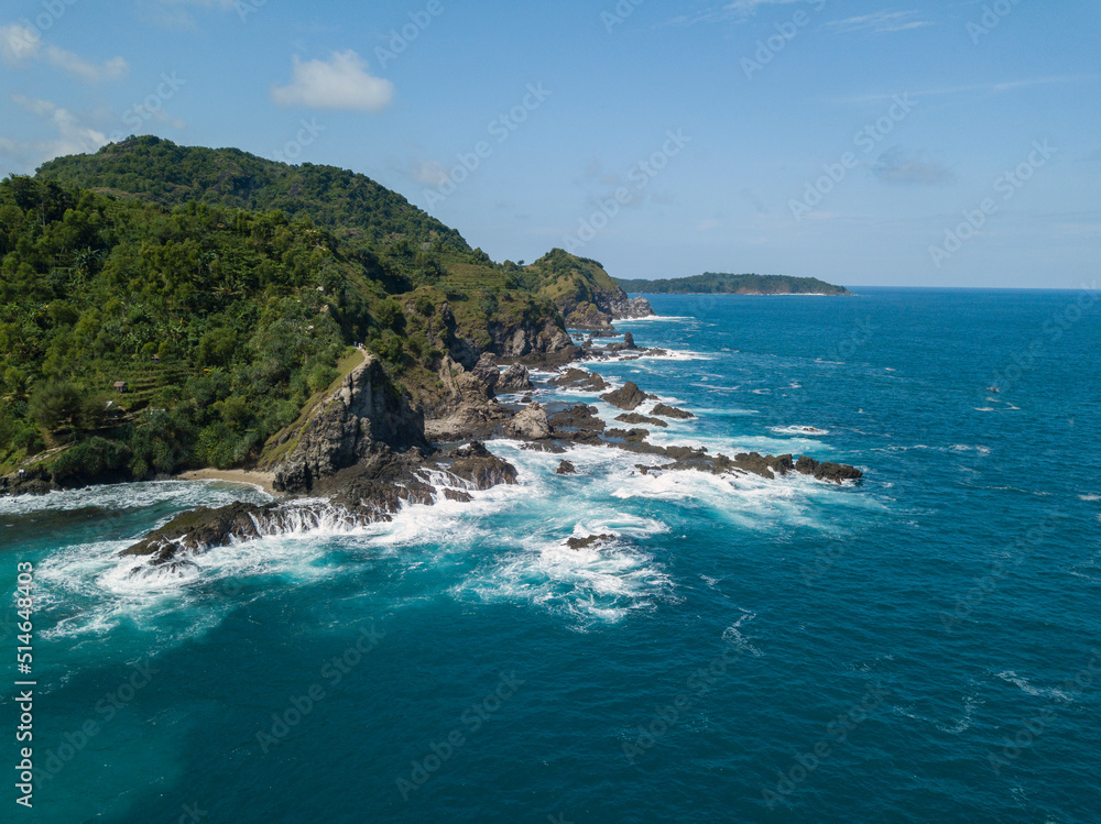Cliffs overgrown by trees that are directly adjacent to the sea. There are rocks that are washed away by the waves with blue sky. The sea water looks blue. Pengilon Hill, Yogyakarta, Indonesia