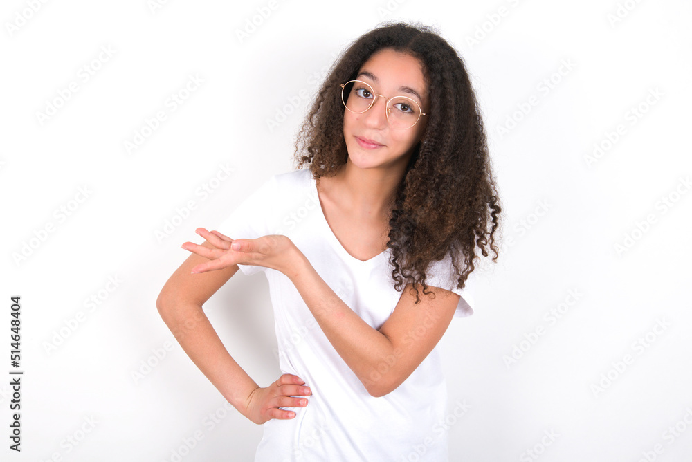 young beautiful girl with afro hairstyle wearing white t-shirt over white wall says: wow how exciting it is, has amazed expression, shows something on blank space with palm. Advertisement concept. young beautiful girl with afro hairstyle wearing white t-shirt over white wall says: wow how exciting it is, has amazed expression, shows something on blank space with palm. Advertisement concept.