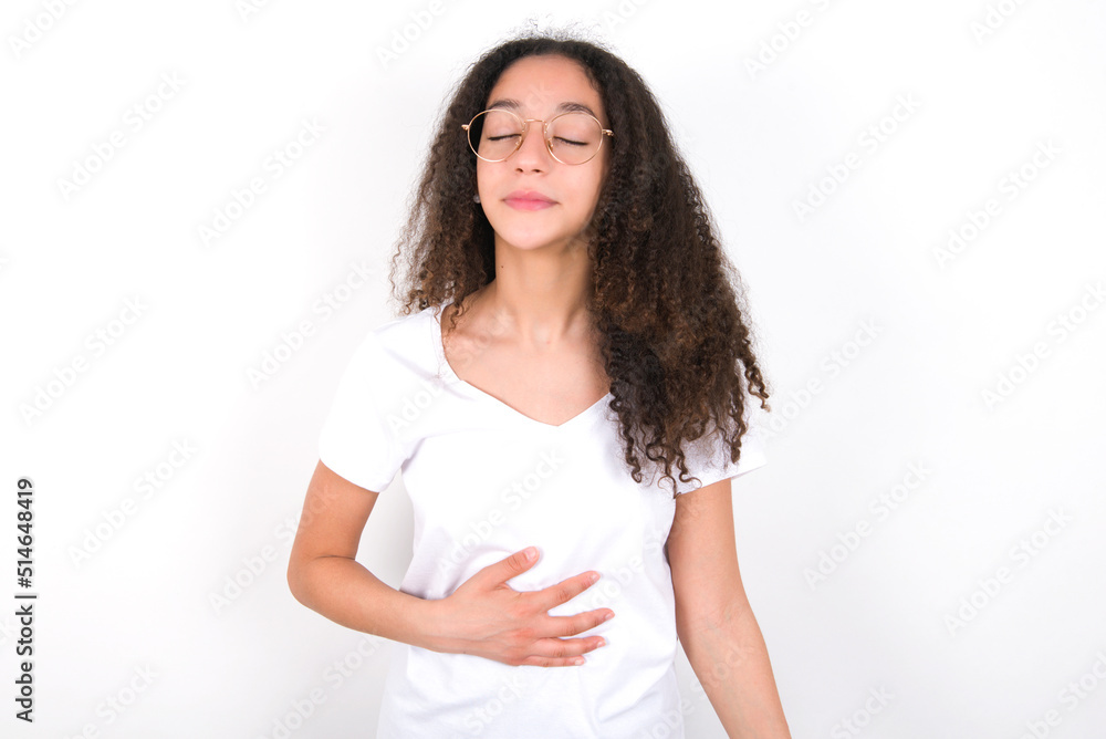 Satisfied smiling young beautiful girl with afro hairstyle wearing white, keeps hands on belly, being in good mood after eating delicious supper, demonstrates she is full. Pleasant feeling in stomach. Satisfied smiling young beautiful girl with afro hairstyle wearing white, keeps hands on belly, being in good mood after eating delicious supper, demonstrates she is full. Pleasant feeling in stomach.
