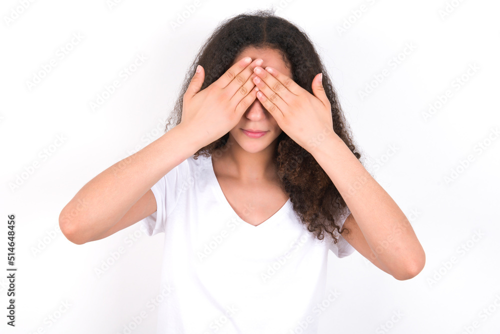 young beautiful girl with afro hairstyle wearing white t-shirt over white wall covering eyes with both hands, doesn't want to see anything or feeling ashamed. Human feelings reactions. young beautiful girl with afro hairstyle wearing white t-shirt over white wall covering eyes with both hands, doesn't want to see anything or feeling ashamed. Human feelings reactions.