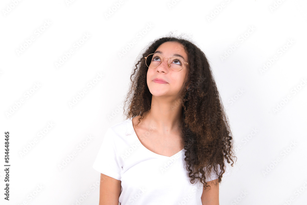 Portrait of mysterious young beautiful girl with afro hairstyle wearing white t-shirt over white wall looking up with enigmatic smile. Advertisement concept. Portrait of mysterious young beautiful girl with afro hairstyle wearing white t-shirt over white wall looking up with enigmatic smile. Advertisement concept.