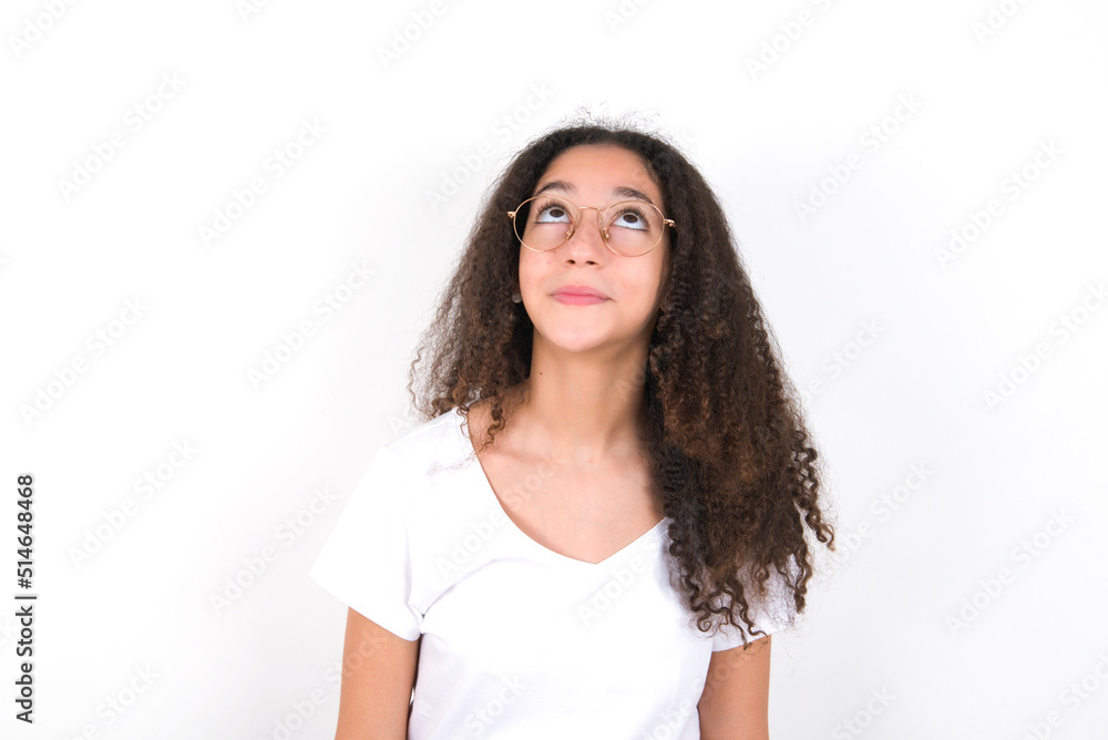 young beautiful girl with afro hairstyle wearing white t-shirt over white wall looking up as he sees something strange. young beautiful girl with afro hairstyle wearing white t-shirt over white wall looking up as he sees something strange.