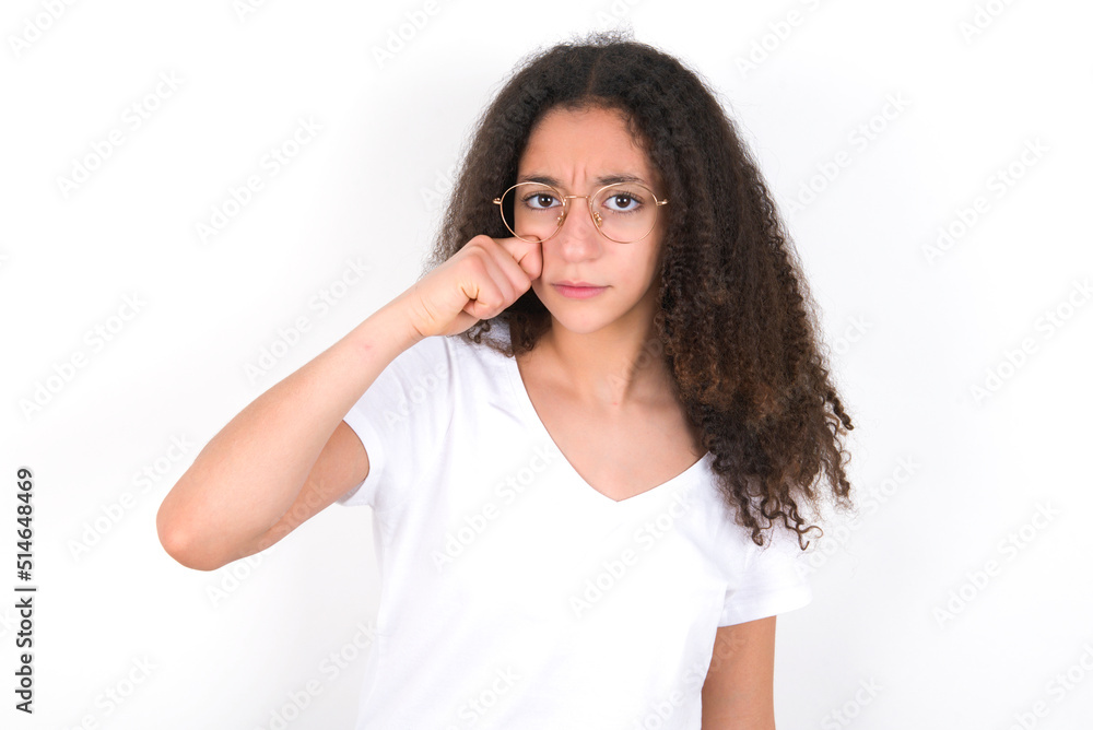 Unhappy young beautiful girl with afro hairstyle wearing white t-shirt over white wall crying while posing at camera whipping tears with hand. Unhappy young beautiful girl with afro hairstyle wearing white t-shirt over white wall crying while posing at camera whipping tears with hand.