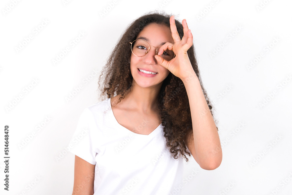 young beautiful girl with afro hairstyle wearing white t-shirt over white wall with happy face smiling doing ok sign with hand on eye looking through finger. young beautiful girl with afro hairstyle wearing white t-shirt over white wall with happy face smiling doing ok sign with hand on eye looking through finger.