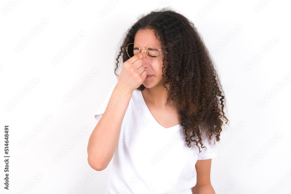 Very upset, young beautiful girl with afro hairstyle wearing white t-shirt over white wall touching nose between closed eyes, wants to cry, having stressful relationship or having troubles with work Very upset, young beautiful girl with afro hairstyle wearing white t-shirt over white wall touching nose between closed eyes, wants to cry, having stressful relationship or having troubles with work