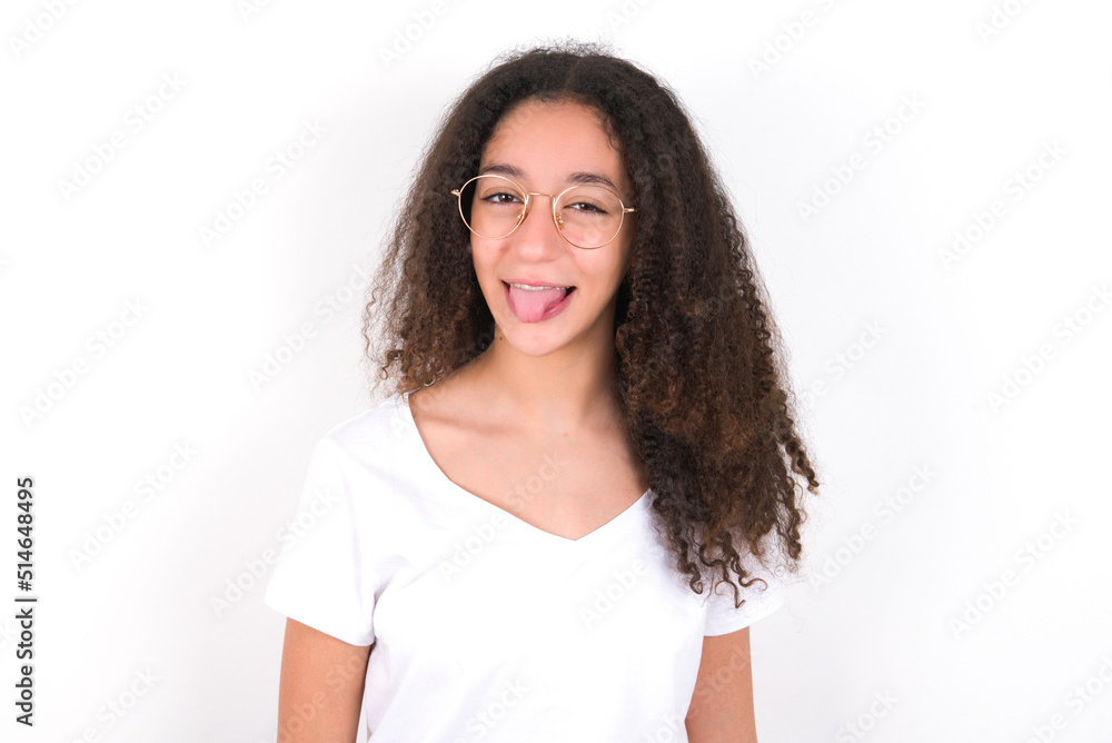 young beautiful girl with afro hairstyle wearing white t-shirt over white wall with happy and funny face smiling and showing tongue. young beautiful girl with afro hairstyle wearing white t-shirt over white wall with happy and funny face smiling and showing tongue.