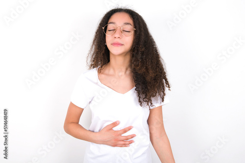 Satisfied smiling young beautiful girl with afro hairstyle wearing white, keeps hands on belly, being in good mood after eating delicious supper, demonstrates she is full. Pleasant feeling in stomach. © Jihan
