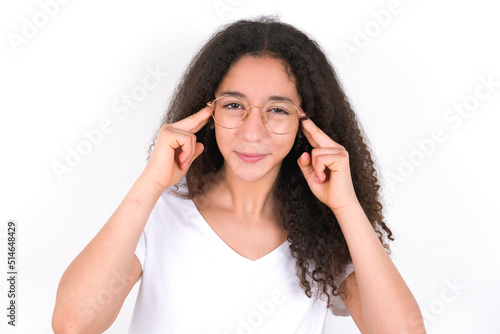 young beautiful girl with afro hairstyle wearing white t-shirt over white wall concentrating hard on an idea with a serious look, thinking with both index fingers pointing to forehead.