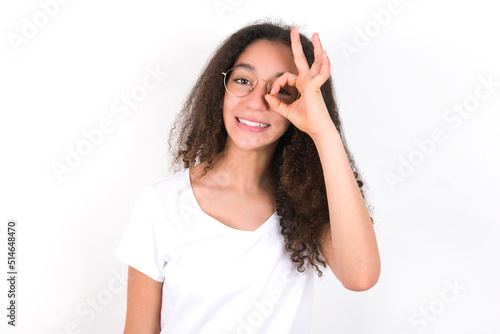 young beautiful girl with afro hairstyle wearing white t-shirt over white wall with happy face smiling doing ok sign with hand on eye looking through finger.