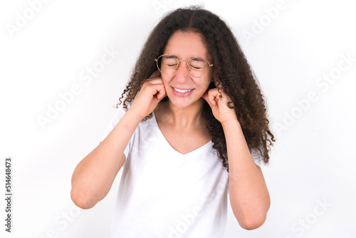 Stop making this annoying sound! Unhappy stressed out young beautiful girl with afro hairstyle wearing white t-shirt over whit making worry face, plugging ears with fingers, irritated with loud noise.