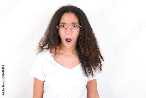 Expressive facial expressions. Shocked stupefied young beautiful girl with afro hairstyle wearing white t-shirt over white wall, keeps jaw dropped feels stunned from what he sees aside. © Jihan