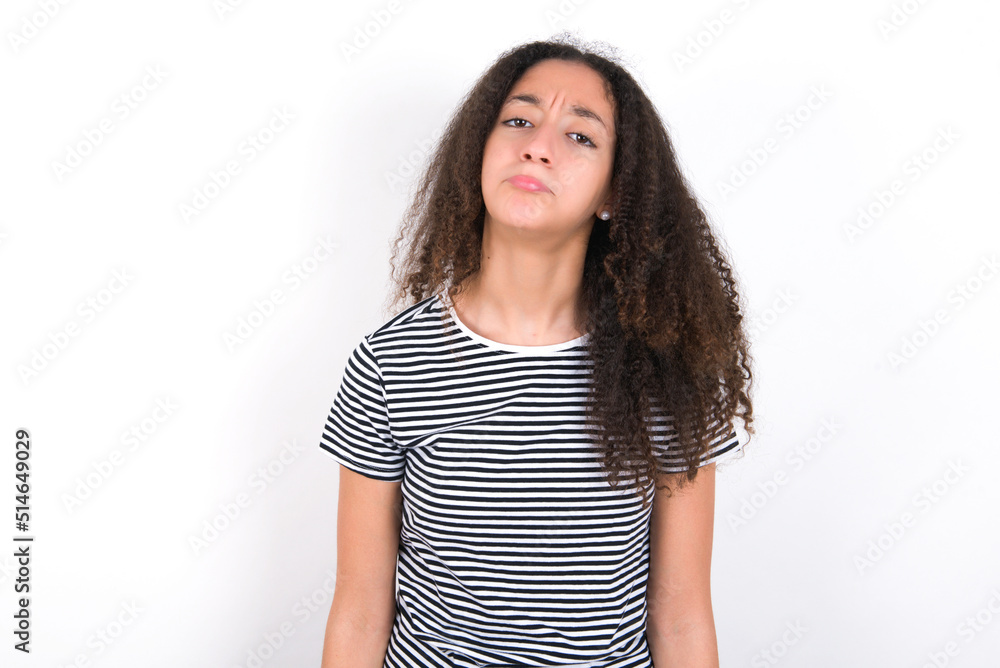 Gloomy, bored young beautiful girl with afro hairstyle wearing striped t-shirt over white wall frowns face looking up, being upset with so much talking hands down, feels tired and wants to leave. Gloomy, bored young beautiful girl with afro hairstyle wearing striped t-shirt over white wall frowns face looking up, being upset with so much talking hands down, feels tired and wants to leave.