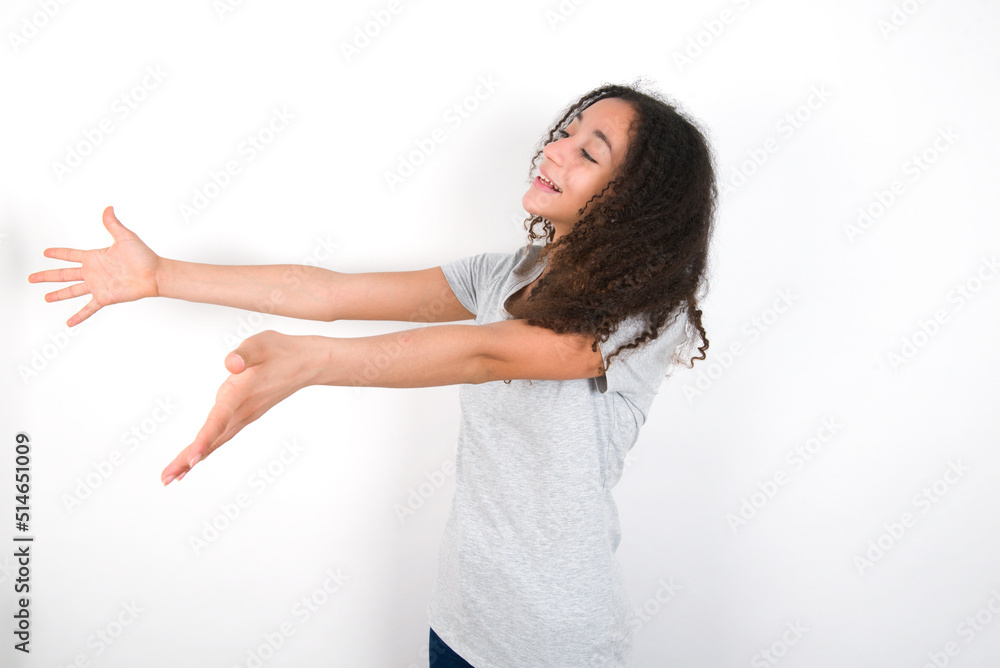 Funny astonished young beautiful girl with afro hairstyle wearing grey t-shirt over white wall look empty space with arms opened ready to catch something. Funny astonished young beautiful girl with afro hairstyle wearing grey t-shirt over white wall look empty space with arms opened ready to catch something.
