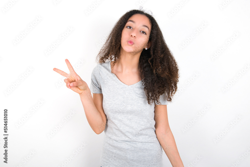 young beautiful girl with afro hairstyle wearing grey t-shirt over white wall makes peace gesture keeps lips folded shows v sign. Body language concept young beautiful girl with afro hairstyle wearing grey t-shirt over white wall makes peace gesture keeps lips folded shows v sign. Body language concept
