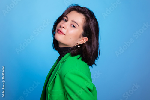 Confident young woman smiling, looking at camera dressed in green blazer isolated on blue background. Studio portrait of successful friendly female with short and dark hair