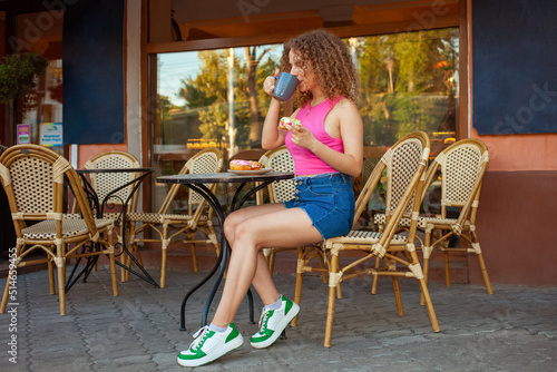 young attractive curly woman sits at table in cafe on summer terrace. Colorful delicious donuts, sweet pastries. Good mood, smiling. Rest, snack break. casual stylish clothes