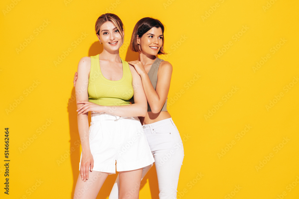 Two young beautiful smiling brunette hipster female in trendy summer shorts clothes. Sexy carefree women posing near yellow wall. Positive models having fun. Cheerful and happy Two young beautiful smiling brunette hipster female in trendy summer shorts clothes. Sexy carefree women posing near yellow wall. Positive models having fun. Cheerful and happy