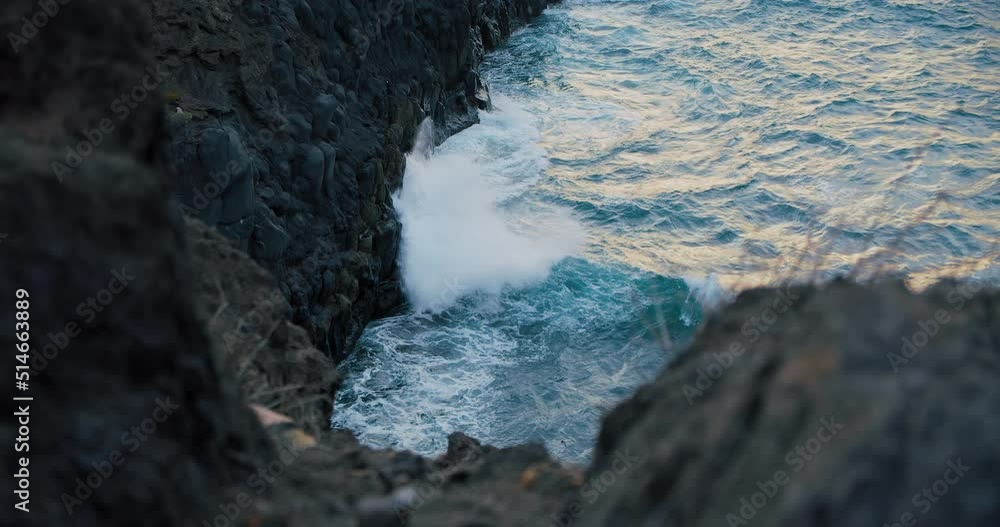 Big waves crash into the black volcanic rocks. Cinematic sunset shot of a deep blue ocean swell violently splashing over the rocky coast of an exotic island. Big waves crash into the black volcanic rocks. Cinematic sunset shot of a deep blue ocean swell violently splashing over the rocky coast of an exotic island.