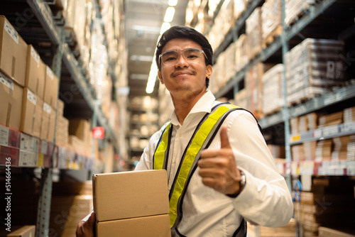 Asian man holds a cardboard box between shelves in a large warehouse. Picking and packing concept Warehouse and workers transport concept.