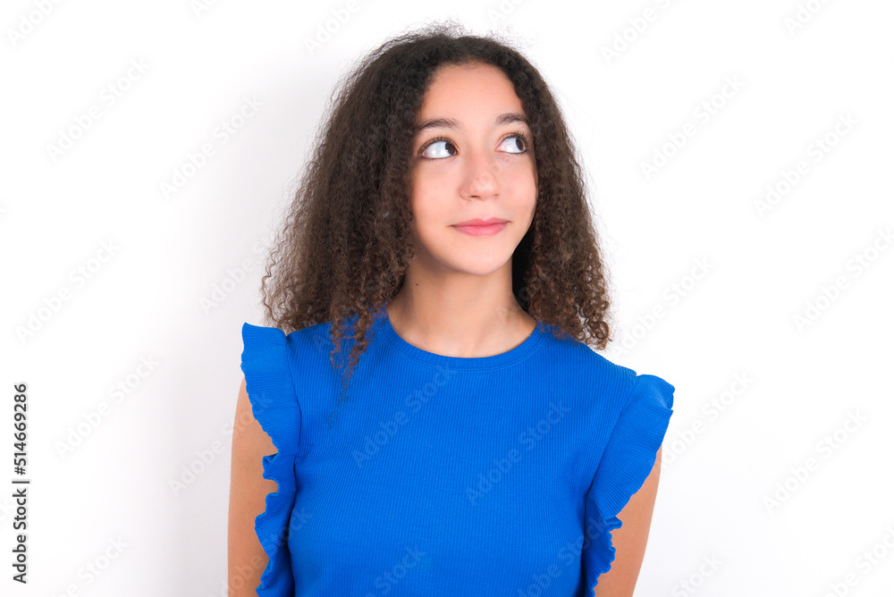 Teenager girl with afro hairstyle wearing blue T-shirt over white wall looking aside into empty space thoughtful Teenager girl with afro hairstyle wearing blue T-shirt over white wall looking aside into empty space thoughtful