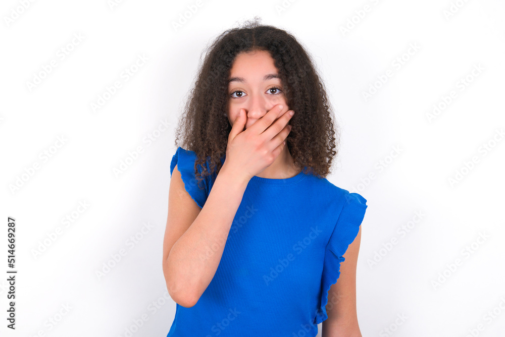 Emotional Teenager girl with afro hairstyle wearing blue T-shirt over white wall gasps from astonishment, covers opened mouth with palm, looks shocked at camera. Reaction concept Emotional Teenager girl with afro hairstyle wearing blue T-shirt over white wall gasps from astonishment, covers opened mouth with palm, looks shocked at camera. Reaction concept