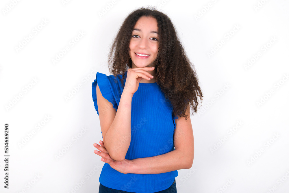 Optimistic Teenager girl with afro hairstyle wearing blue T-shirt over white wall keeps hands partly crossed and hand under chin, looks at camera with pleasure. Happy emotions concept. Optimistic Teenager girl with afro hairstyle wearing blue T-shirt over white wall keeps hands partly crossed and hand under chin, looks at camera with pleasure. Happy emotions concept.