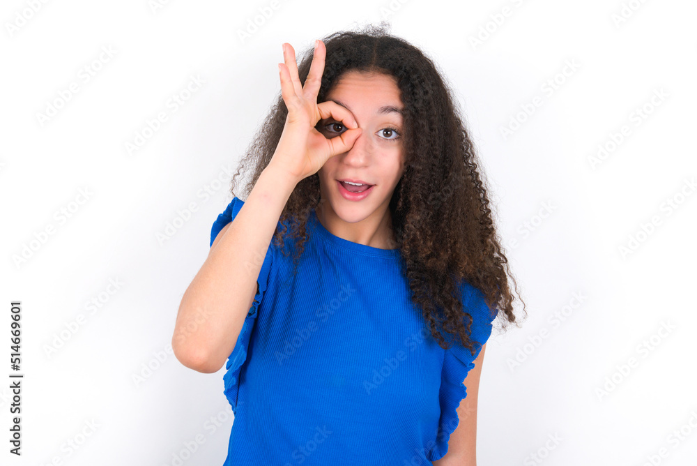 Teenager girl with afro hairstyle wearing blue T-shirt over white wall doing ok gesture shocked with surprised face, eye looking through fingers. Unbelieving expression. Teenager girl with afro hairstyle wearing blue T-shirt over white wall doing ok gesture shocked with surprised face, eye looking through fingers. Unbelieving expression.