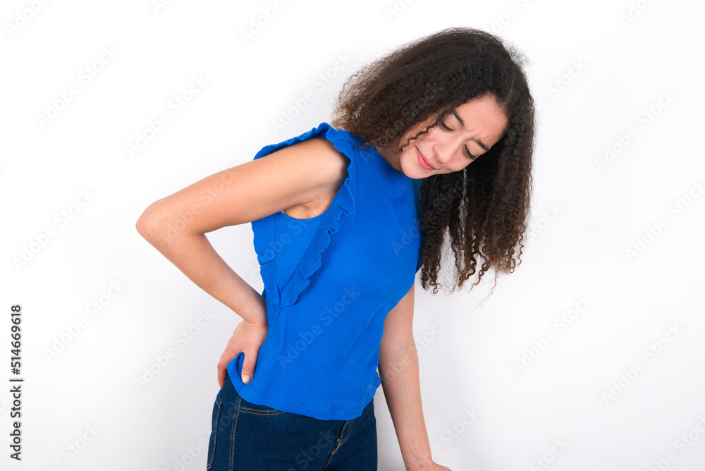 Teenager girl with afro hairstyle wearing blue T-shirt over white wall Suffering of backache, touching back with hand, muscular pain Teenager girl with afro hairstyle wearing blue T-shirt over white wall Suffering of backache, touching back with hand, muscular pain