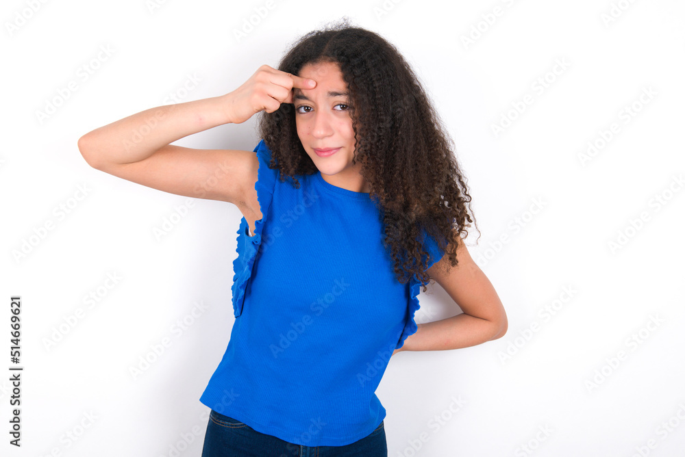 Teenager girl with afro hairstyle wearing blue T-shirt over white wall pointing unhappy at pimple on forehead, blackhead infection. Skincare concept. Teenager girl with afro hairstyle wearing blue T-shirt over white wall pointing unhappy at pimple on forehead, blackhead infection. Skincare concept.