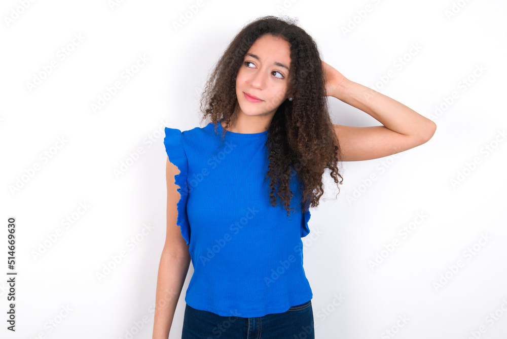 Teenager girl with afro hairstyle wearing blue T-shirt over white wall confuse and wonder about question. Uncertain with doubt, thinking with hand on head. Pensive concept. Teenager girl with afro hairstyle wearing blue T-shirt over white wall confuse and wonder about question. Uncertain with doubt, thinking with hand on head. Pensive concept.