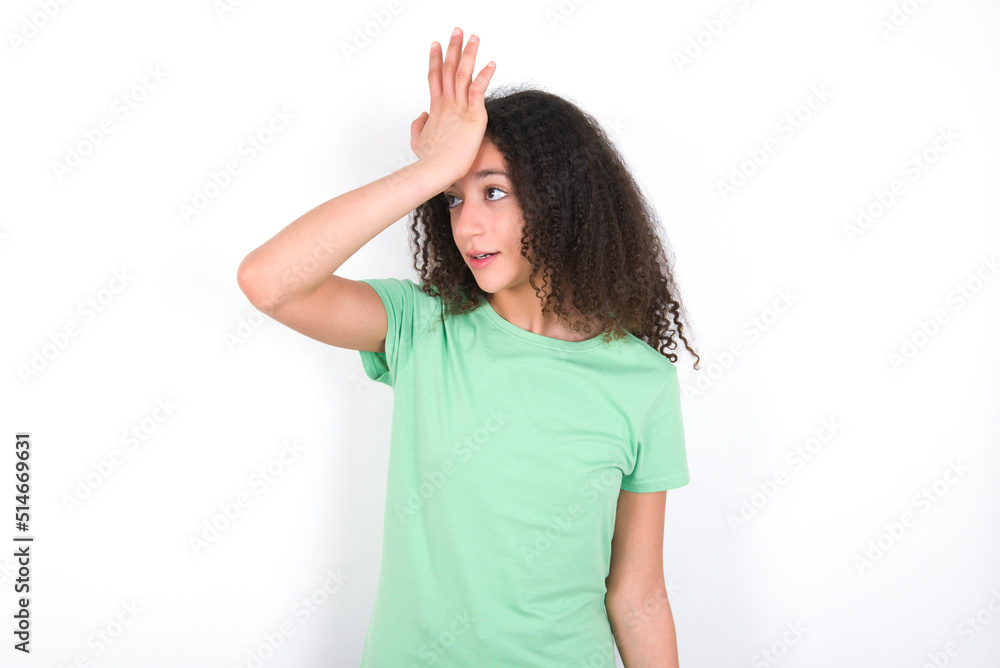 Teenager girl with afro hairstyle wearing green T-shirt over white wall surprised with hand on head for mistake, remember error. Forgot, bad memory concept. Teenager girl with afro hairstyle wearing green T-shirt over white wall surprised with hand on head for mistake, remember error. Forgot, bad memory concept.