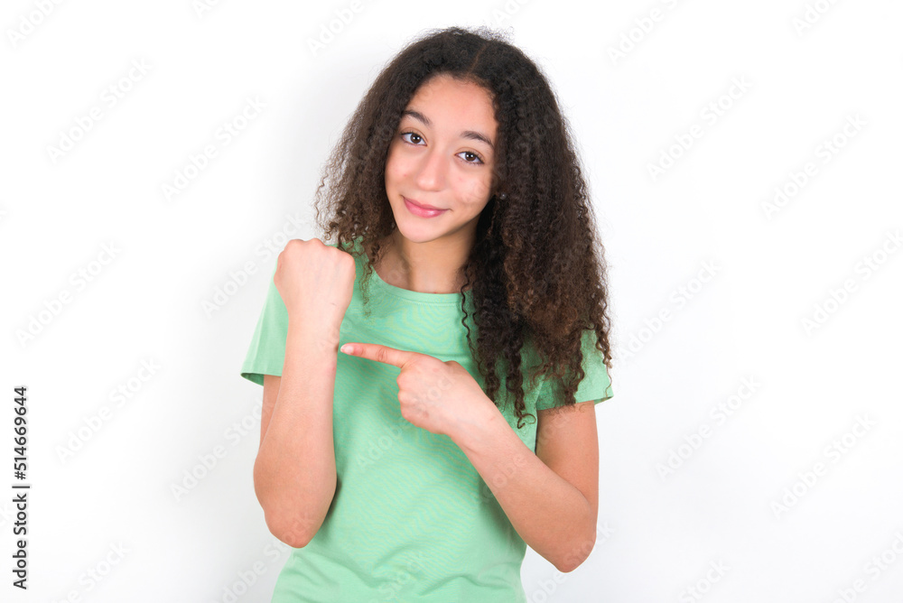 Teenager girl with afro hairstyle wearing green T-shirt over white wall In hurry pointing to wrist watch, impatience, looking at the camera with relaxed expression Teenager girl with afro hairstyle wearing green T-shirt over white wall In hurry pointing to wrist watch, impatience, looking at the camera with relaxed expression