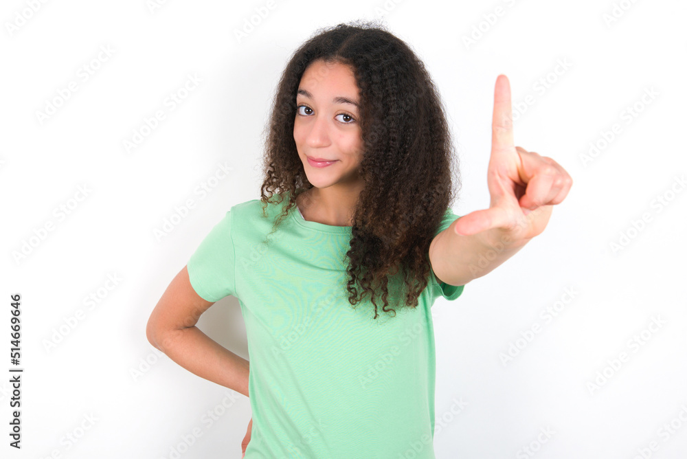 Teenager girl with afro hairstyle wearing green T-shirt over white wall making fun of people with fingers on forehead doing loser gesture mocking and insulting. Teenager girl with afro hairstyle wearing green T-shirt over white wall making fun of people with fingers on forehead doing loser gesture mocking and insulting.