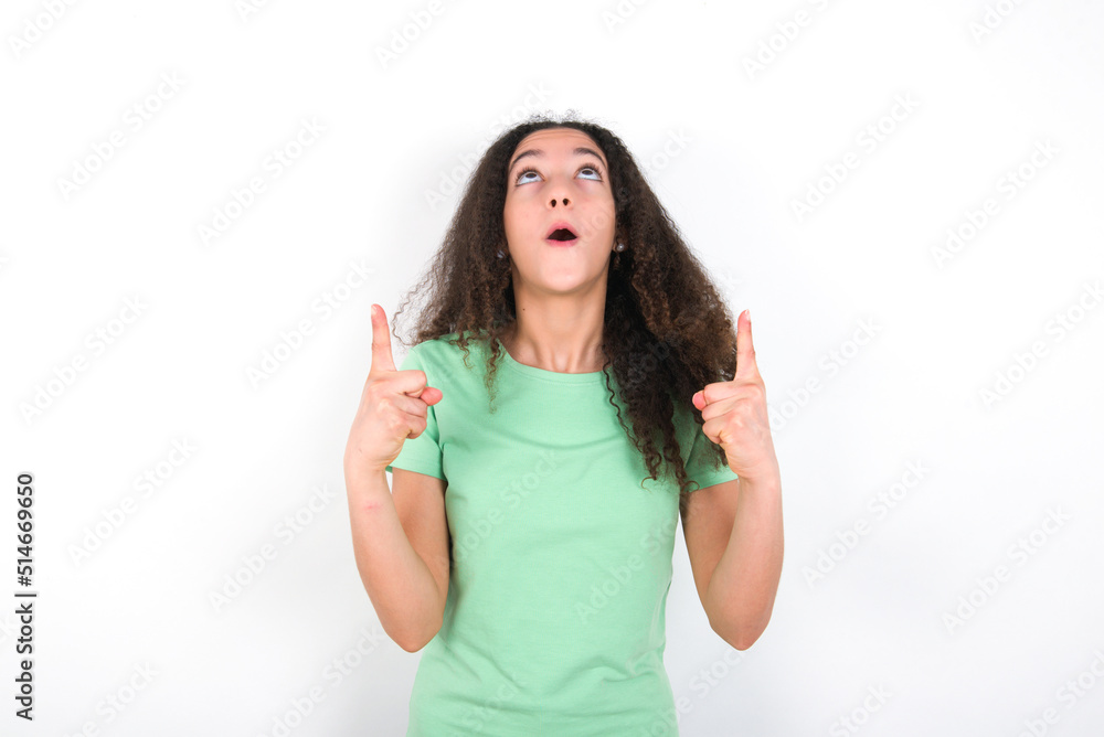 Teenager girl with afro hairstyle wearing green T-shirt over white wall amazed and surprised looking up and pointing with fingers and raised arms. Teenager girl with afro hairstyle wearing green T-shirt over white wall amazed and surprised looking up and pointing with fingers and raised arms.