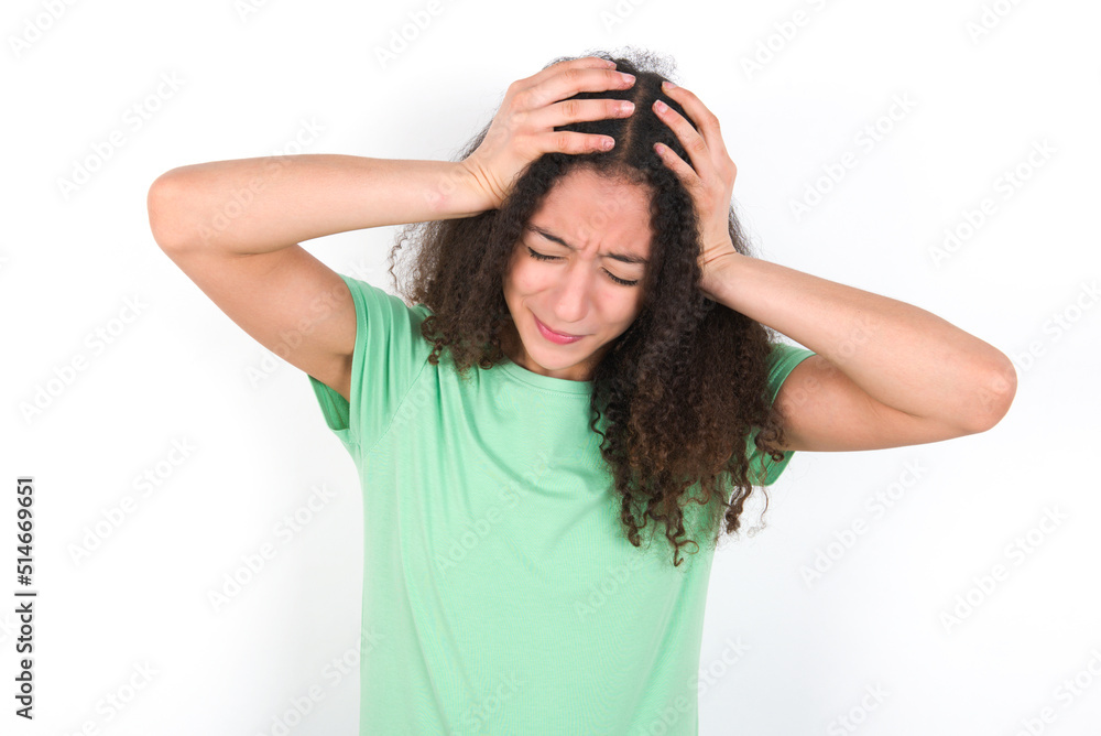 Teenager girl with afro hairstyle wearing green T-shirt over white wall suffering from strong headache desperate and stressed because of overwork. Depression and pain concept. Teenager girl with afro hairstyle wearing green T-shirt over white wall suffering from strong headache desperate and stressed because of overwork. Depression and pain concept.