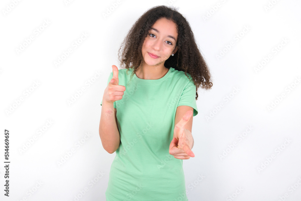 Teenager girl with afro hairstyle wearing green T-shirt over white wall pointing fingers to camera with happy and funny face. Good energy and vibes. Teenager girl with afro hairstyle wearing green T-shirt over white wall pointing fingers to camera with happy and funny face. Good energy and vibes.