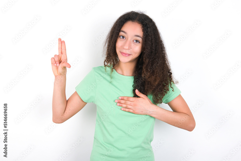 Teenager girl with afro hairstyle wearing green T-shirt over white wall smiling swearing with hand on chest and fingers up, making a loyalty promise oath. Teenager girl with afro hairstyle wearing green T-shirt over white wall smiling swearing with hand on chest and fingers up, making a loyalty promise oath.