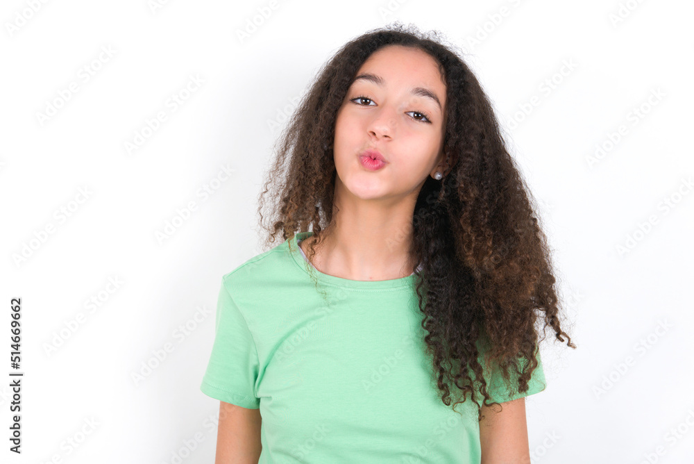 Shot of pleasant looking Teenager girl with afro hairstyle wearing green T-shirt over white wall, pouts lips, looks at camera, Human facial expressions Shot of pleasant looking Teenager girl with afro hairstyle wearing green T-shirt over white wall, pouts lips, looks at camera, Human facial expressions