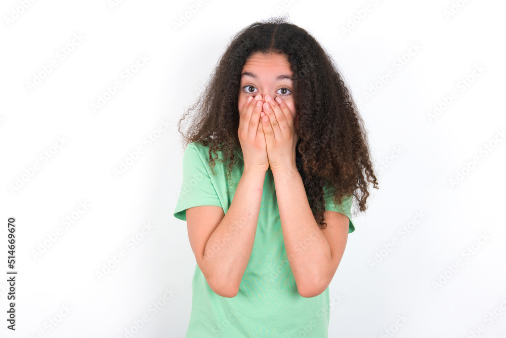 Vivacious Teenager girl with afro hairstyle wearing green T-shirt over white wall, giggles joyfully, covers mouth, has natural laughter, hears positive story or funny anecdote Vivacious Teenager girl with afro hairstyle wearing green T-shirt over white wall, giggles joyfully, covers mouth, has natural laughter, hears positive story or funny anecdote