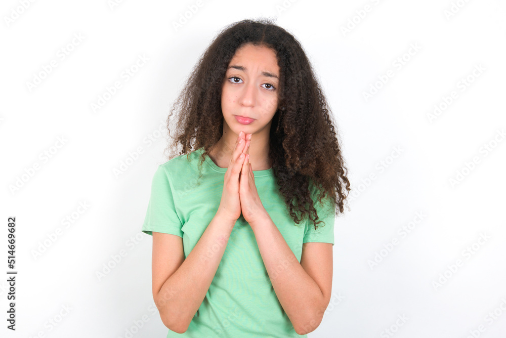 Positive Teenager girl with afro hairstyle wearing green T-shirt over white wall smiles happily, glad to receive pleasant news from interlocutor, keeps palms together, People emotions concept.