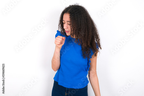 Teenager girl with afro hairstyle wearing blue T-shirt over white wall feeling unwell and coughing as symptom for cold or bronchitis. Healthcare concept.