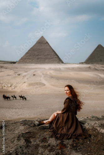 Young redhead tourist girl in brown dress sitting on a stone cliff in Egypt, Cairo - Giza. Pyramids on backround. Copy space