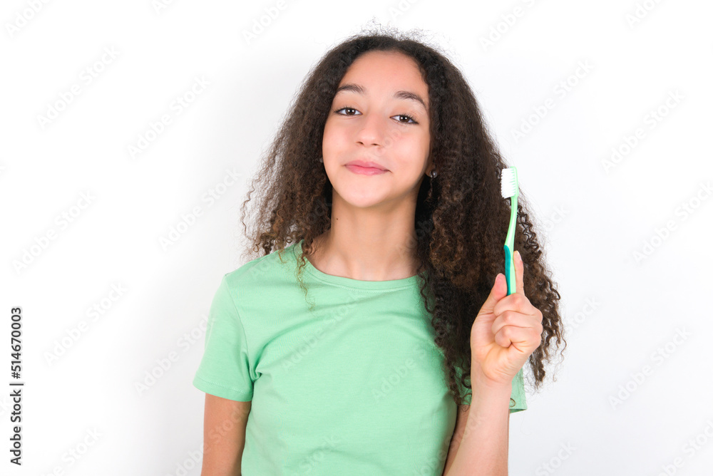 Teenager girl with afro hairstyle wearing green T-shirt over white wall holding a toothbrush and smiling. Dental healthcare concept. Teenager girl with afro hairstyle wearing green T-shirt over white wall holding a toothbrush and smiling. Dental healthcare concept.