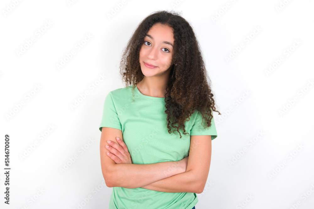 Self confident serious calm Teenager girl with afro hairstyle wearing green T-shirt over white wall stands with arms folded. Shows professional vibe stands in assertive pose. Self confident serious calm Teenager girl with afro hairstyle wearing green T-shirt over white wall stands with arms folded. Shows professional vibe stands in assertive pose.