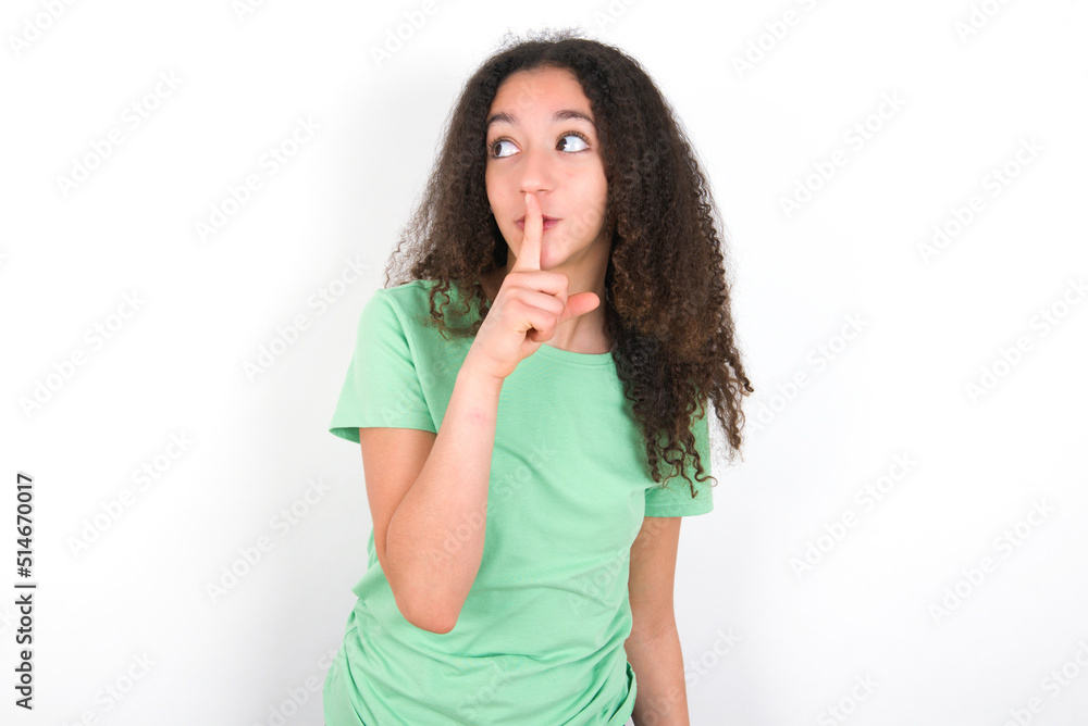 Teenager girl with afro hairstyle wearing green T-shirt over white wall silence gesture keeps index finger to lips makes hush sign. Asks not to share secret Teenager girl with afro hairstyle wearing green T-shirt over white wall silence gesture keeps index finger to lips makes hush sign. Asks not to share secret