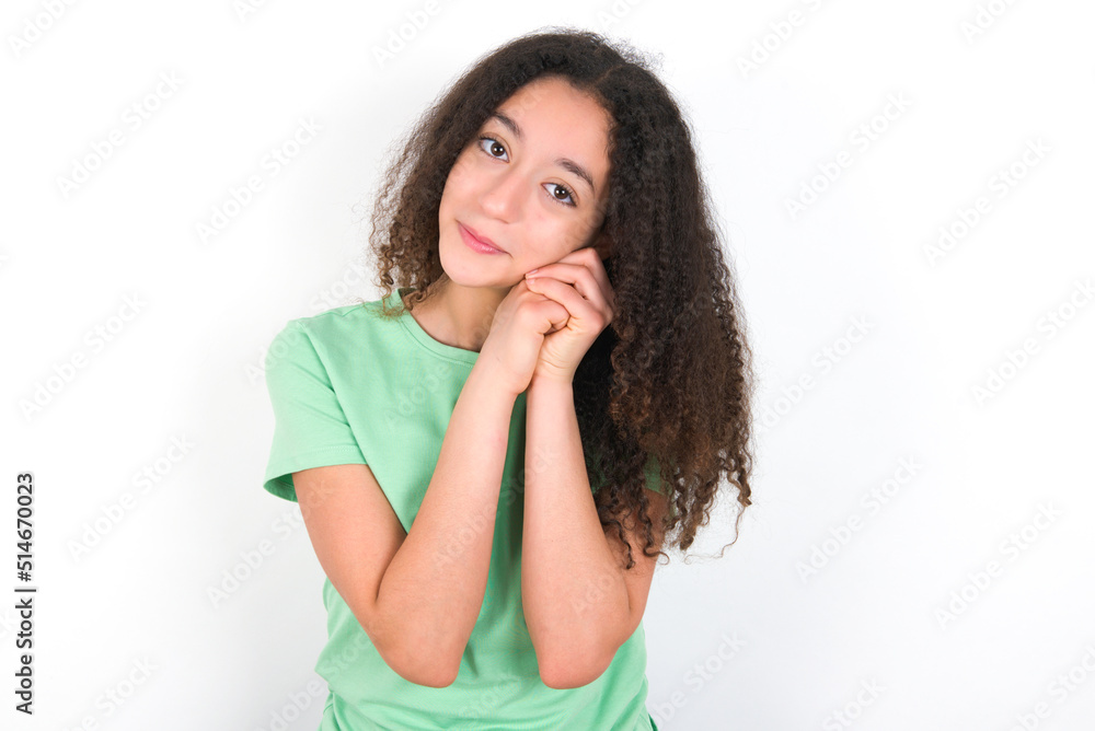 Charming serious Teenager girl with afro hairstyle wearing green T-shirt over white wall  keeps hands near face smiles tenderly at camera