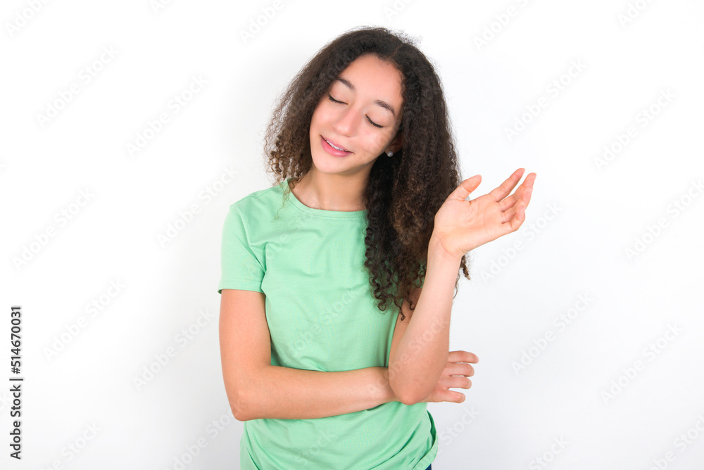 Overjoyed successful Teenager girl with afro hairstyle wearing green T-shirt over white wall raises palm and closes eyes in joy being entertained by friends Overjoyed successful Teenager girl with afro hairstyle wearing green T-shirt over white wall raises palm and closes eyes in joy being entertained by friends