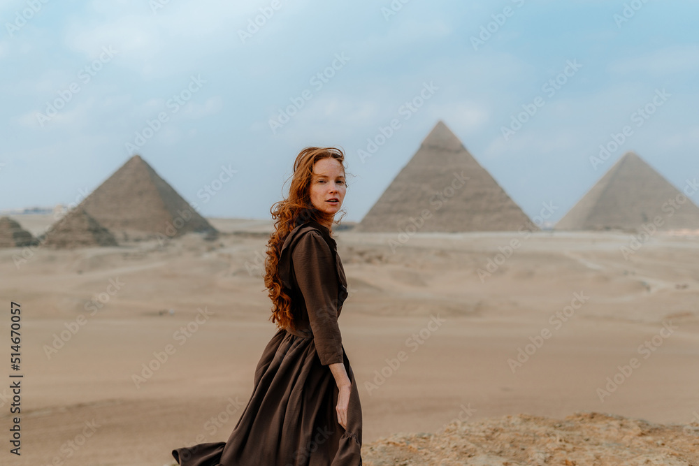 Young redhead tourist girl wearing a brown dress standing on the sand in Egypt, Cairo - Giza. Pyramids on backround. Copy space - obrazy, fototapety, plakaty 