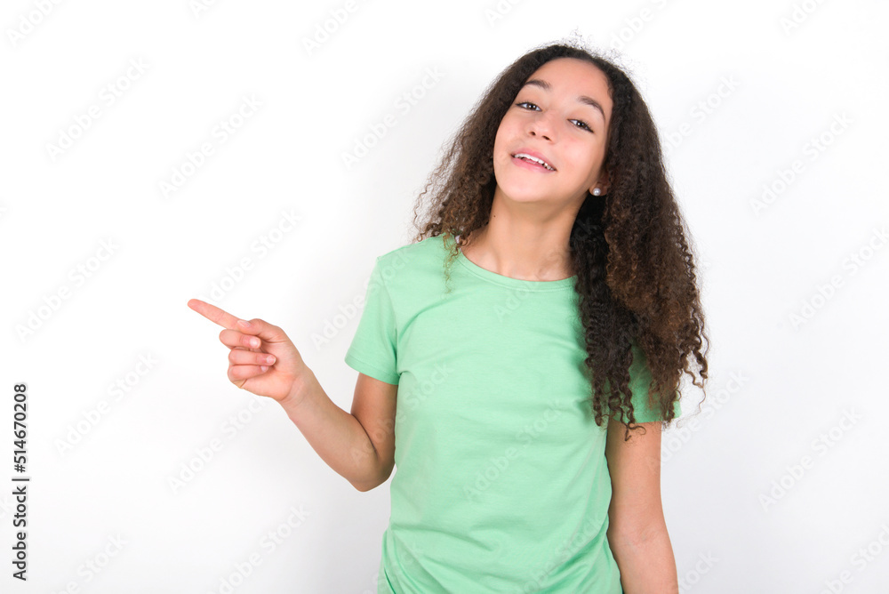 Teenager girl with afro hairstyle wearing green T-shirt over white wall laughs happily points away on blank space demonstrates shopping discount offer, excited by good news or unexpected sale. Teenager girl with afro hairstyle wearing green T-shirt over white wall laughs happily points away on blank space demonstrates shopping discount offer, excited by good news or unexpected sale.