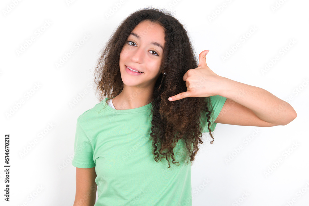 Teenager girl with afro hairstyle wearing green T-shirt over white wall makes phone gesture, says call me back again, has glad expression. Teenager girl with afro hairstyle wearing green T-shirt over white wall makes phone gesture, says call me back again, has glad expression.